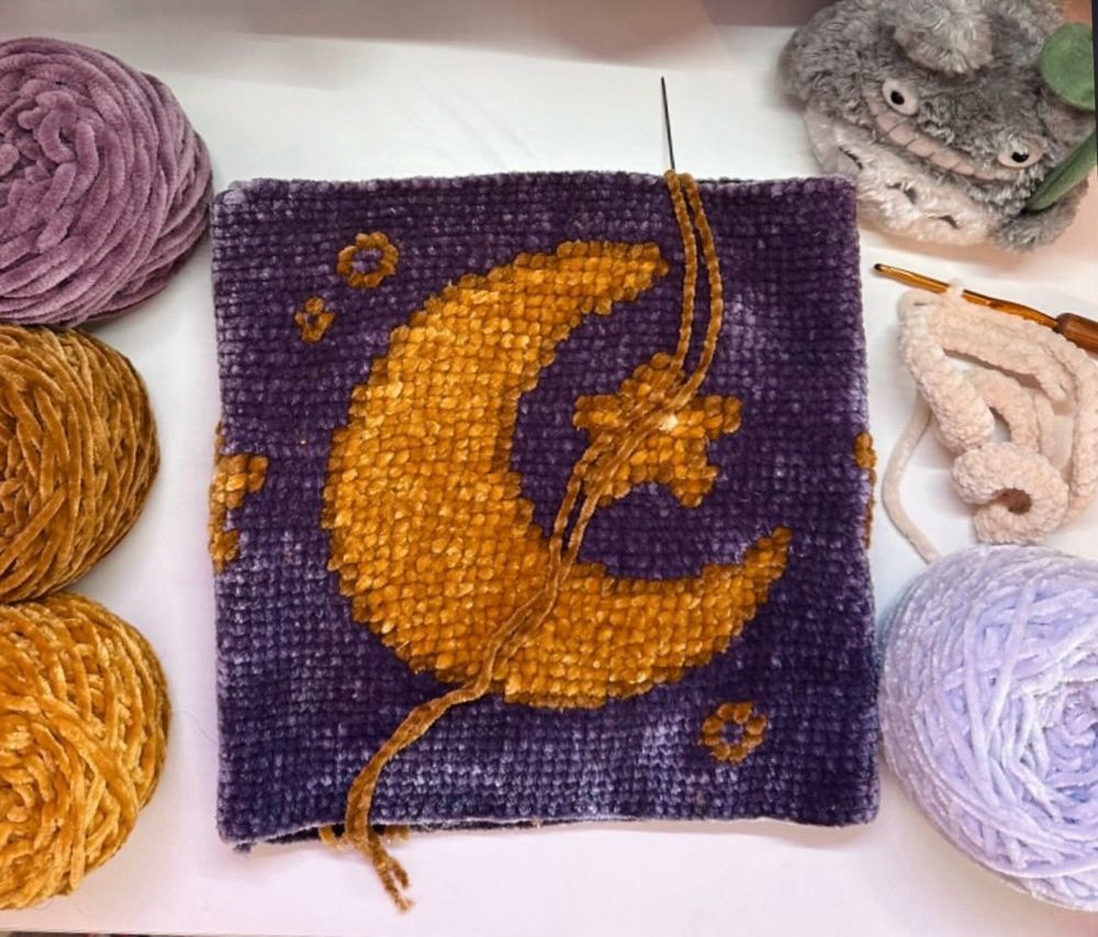  A crochet or cross-stitch panel featuring a golden crescent moon and small stars on a deep purple background. The panel is surrounded by skeins of yarn in complementary shades of gold, lavender, and mauve, along with crochet tools and a soft, plush decorative item in the top right corner. The piece has a celestial, whimsical aesthetic and appears to be a work in progress.