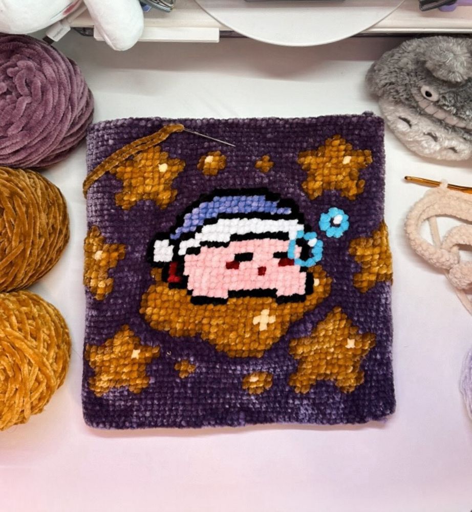 A crochet or cross-stitch panel featuring a pixel-art style design of a sleeping Kirby wearing a blue nightcap, surrounded by golden stars on a purple background. The panel is surrounded by balls of yarn in various colors and crochet hooks, suggesting a work in progress. The scene is cozy and creative, with warm tones and a soft aesthetic.