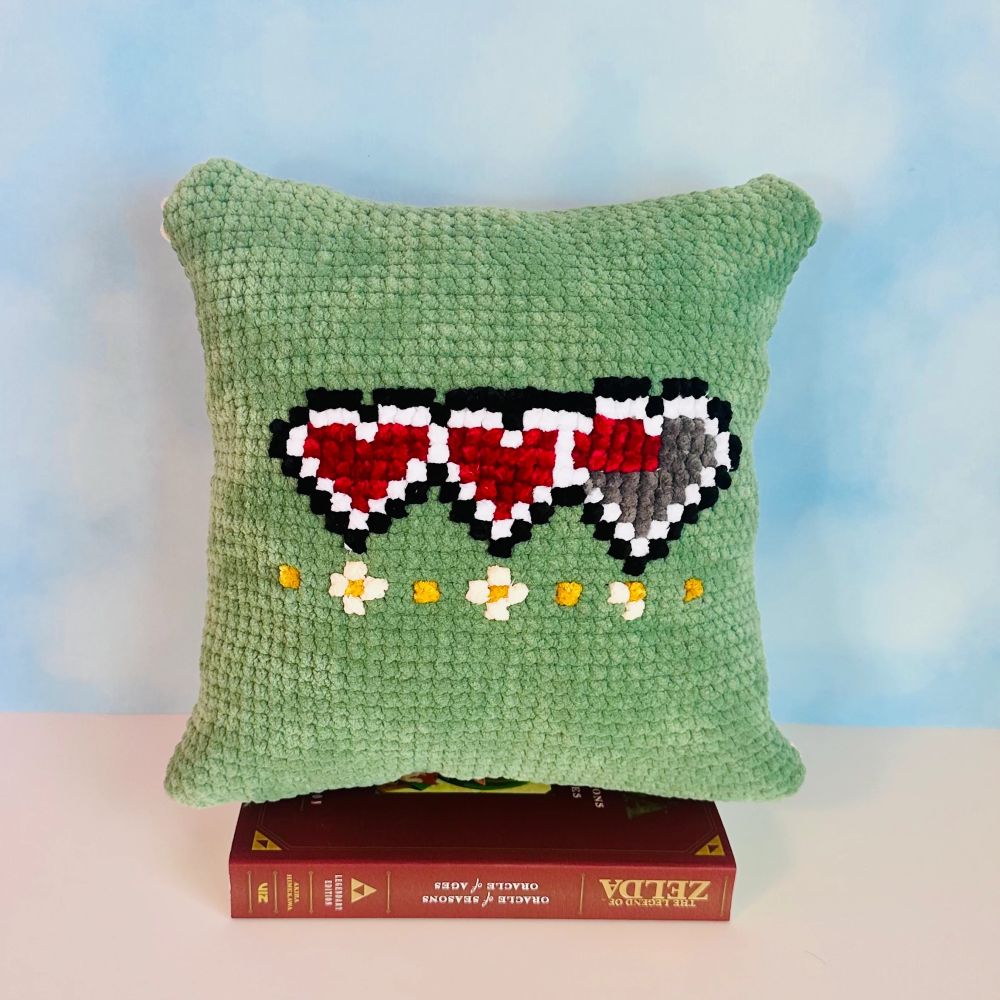  A handcrafted pillow featuring a crochet and cross-stitch design inspired by the Zelda video game series. The front depicts a vibrant yellow Triforce symbol surrounded by a green and white pixelated border, resembling the classic design elements of the game, set against a light green background.