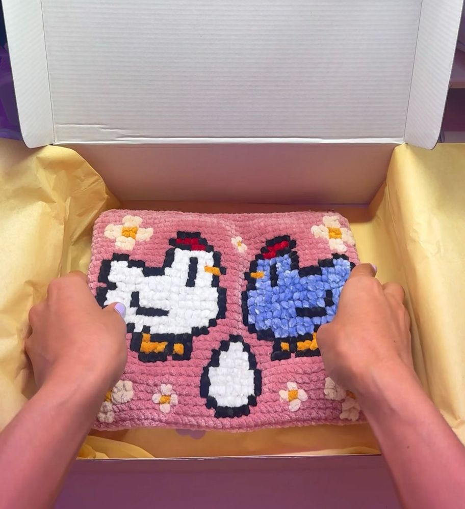 A handmade latch-hook rug featuring a cute design of a white chicken, a blue chicken, and an egg in the center, surrounded by small flowers on a pink background. The rug is displayed inside a gift box lined with yellow tissue paper. Two hands are holding the edges of the rug, emphasizing its vibrant colors and cozy texture, giving the impression of an adorable and heartfelt handmade gift.