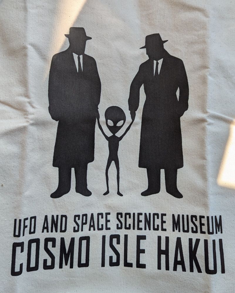 A tote bag with black text and imagery. The image is of two Men In Black type people in suits each holding the hand of a grey alien who is so small it looks like they are holding the hands of a small child. The text reads: UFO AND SPACE SCIENCE MUSEUM COSMO ISLE HAKUI