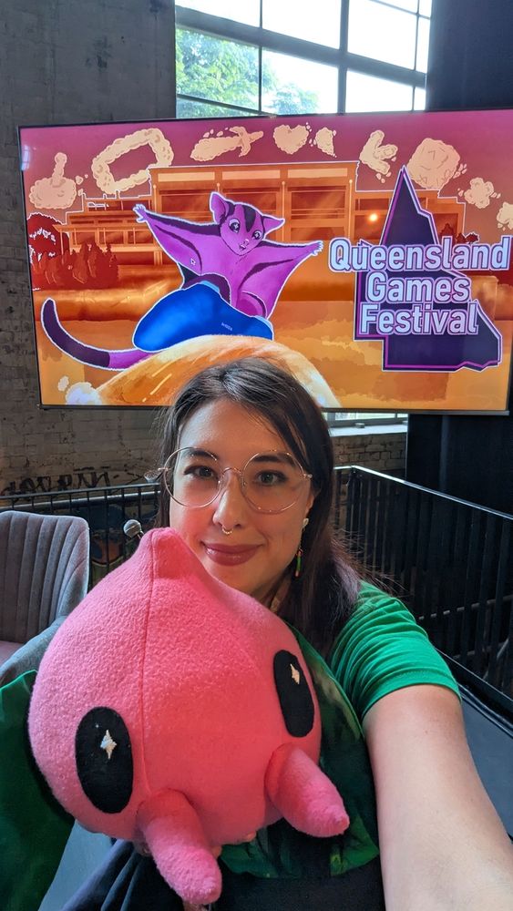 Selfie with Tempopo puppet I made and the Queensland Games Festival 2025 art that is on the TV behind me. Two of my arts in one picture.