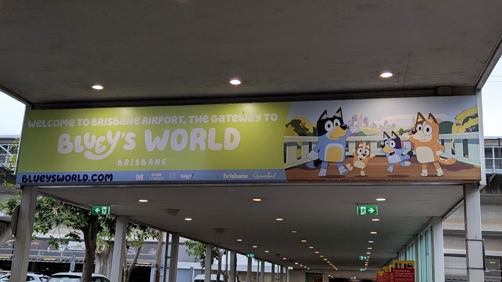 Picture of the banner right outside Brisbane International Airport Arrivals that has a picture of the main four of Bluey's family to the right. To the left the sign reads: WELCOME TO BRISBANE AIRPORT, THE GATEWAY TO BLUEY'S WORLD BRISBANE 