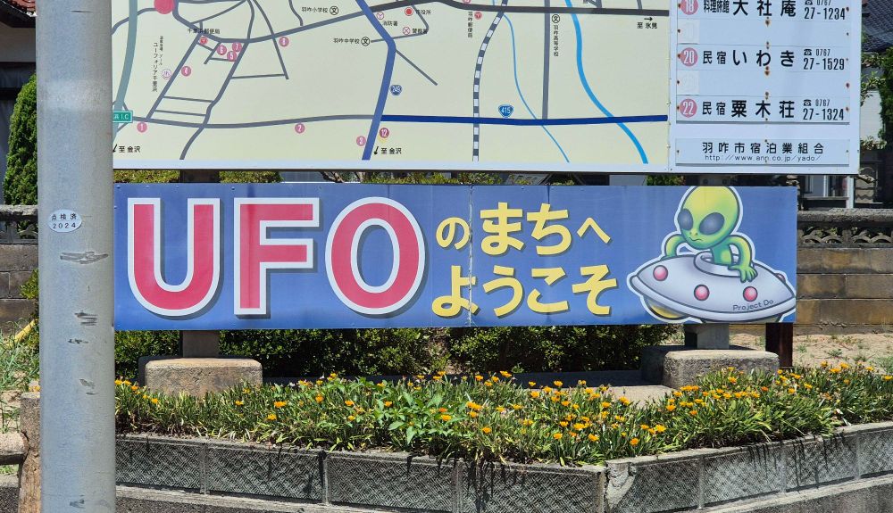 A street sign in English and Japanese that roughly google translates to, 'Welcome to UFO Town'. There is an alien in a spaceship to the right of the words.