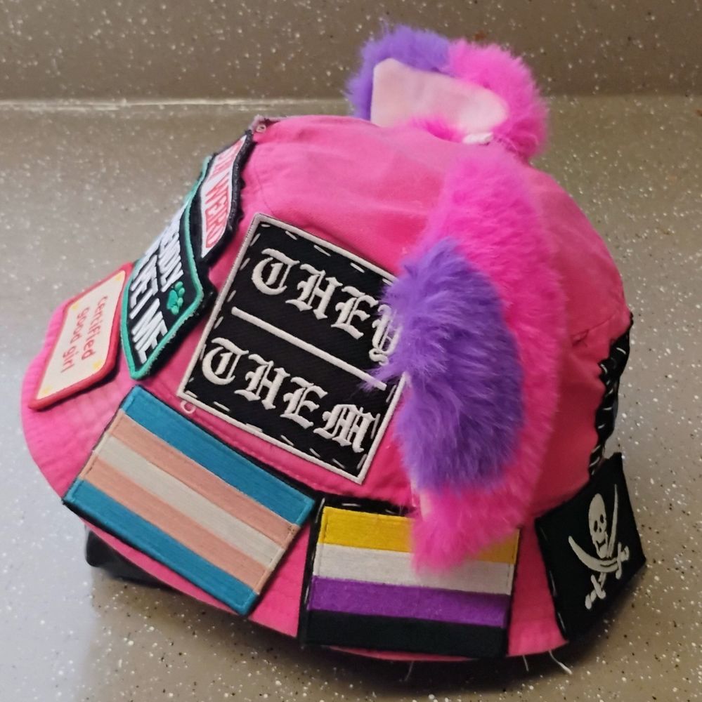 side of my hot pink bucket hat with cat ears and patches sewn on. there's "they/them" and trans and nonbinary pride flags