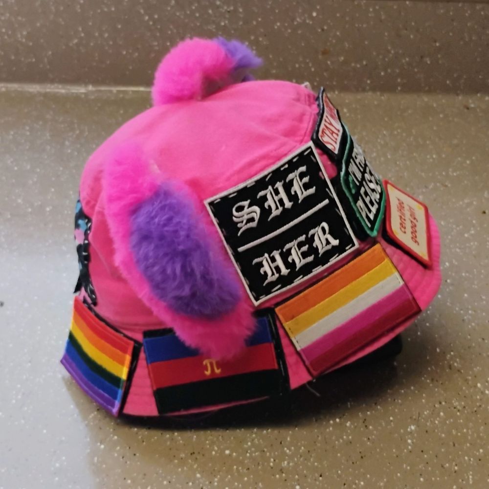 side of my hot pink bucket hat with cat ears and patches sewn on. there's "she/her" and lesbian and polyamory pride flags