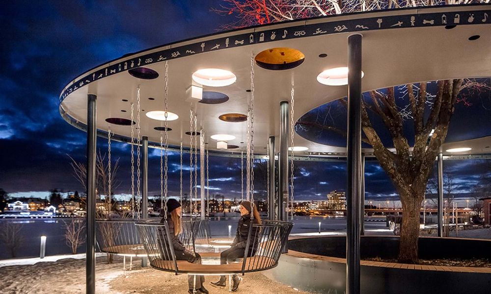 Frizon shelter - a curved space with seats like swings and lovely coloured perspex in the roof and a view of the city of Umeå at dusk. Two teenage girls sit in one of the swing seats having a chat. 