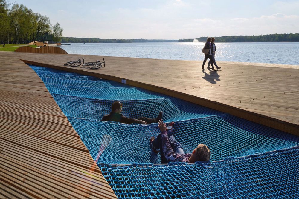 A  long run of blue netted hammocks are set into a wooden boardwalk by a lake; two people are lounging on them; two more walk past on a fine day
