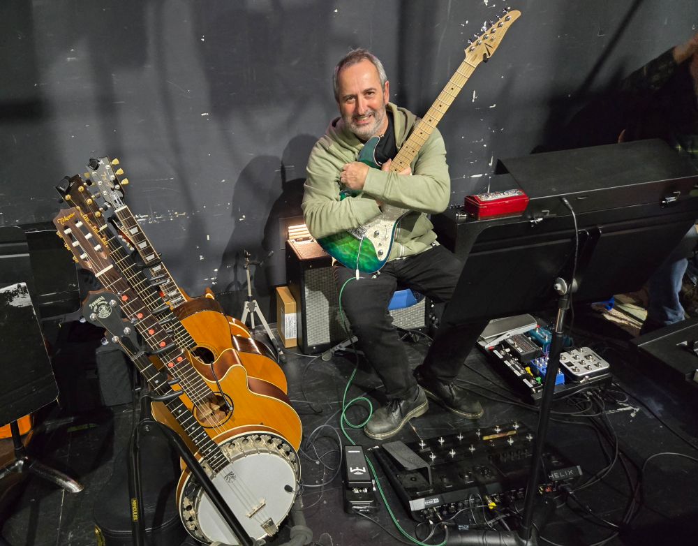 A very happy pit guitar player sitting on a stool surrounded by guitars, amplifiers and pedalboards 