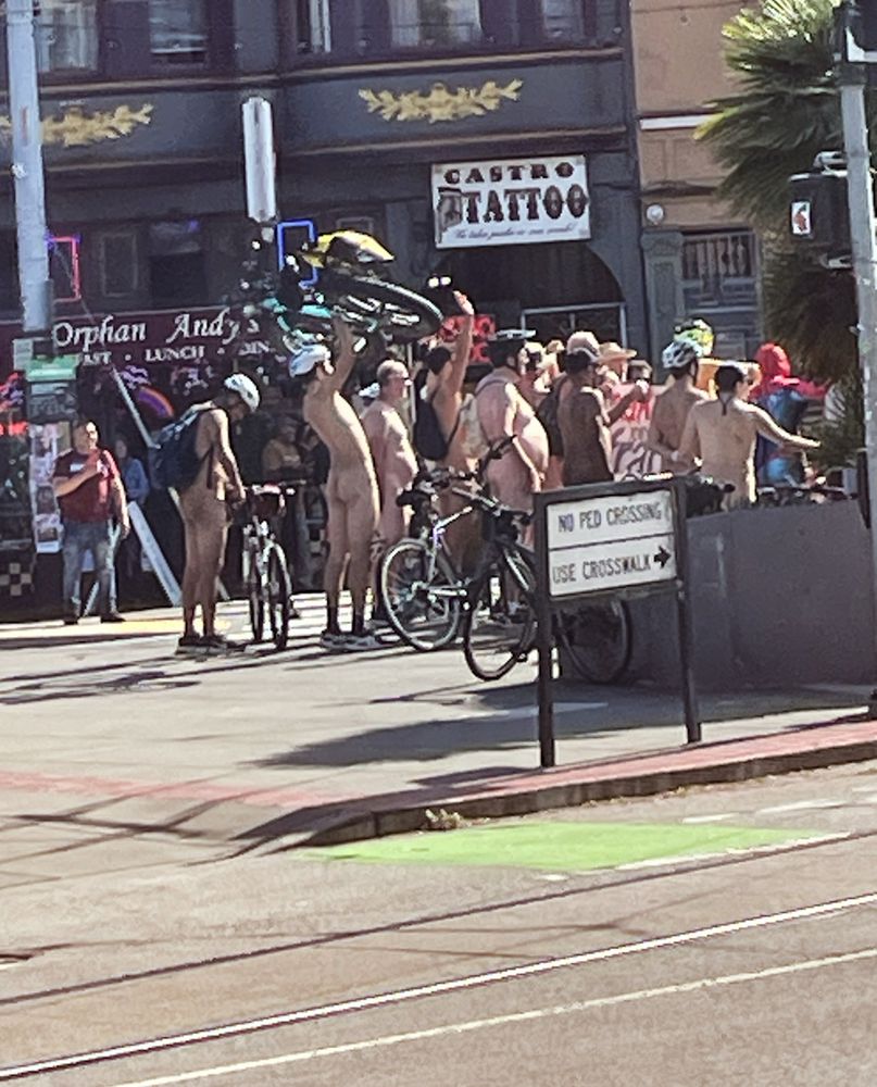 A group of about 30 naked men are posing for a photo by the gas station in Jane Warner Plaza in the Castro, facing Twin Peaks. They’re seen in profile while holding their bikes as the photographer stands out of frame. 