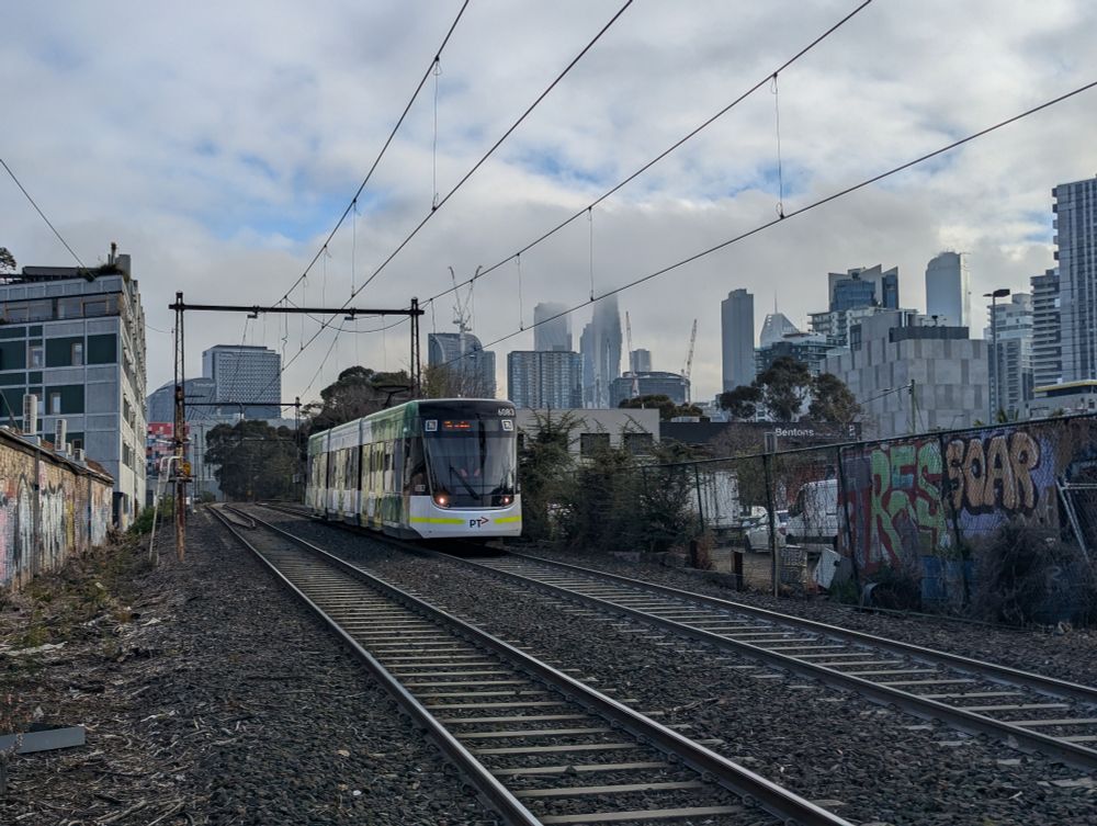 an E class tram running down the former St Kilda railway line with Melbourne's CBD Skyline in the background, partially obscured by clouds