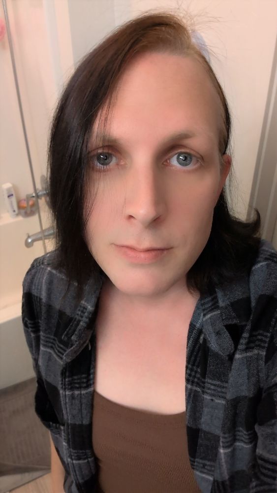 Reyna in her bathroom, her hair is flipped to one side and partially covering her face. She's wearing a grey flannel over a tank top.