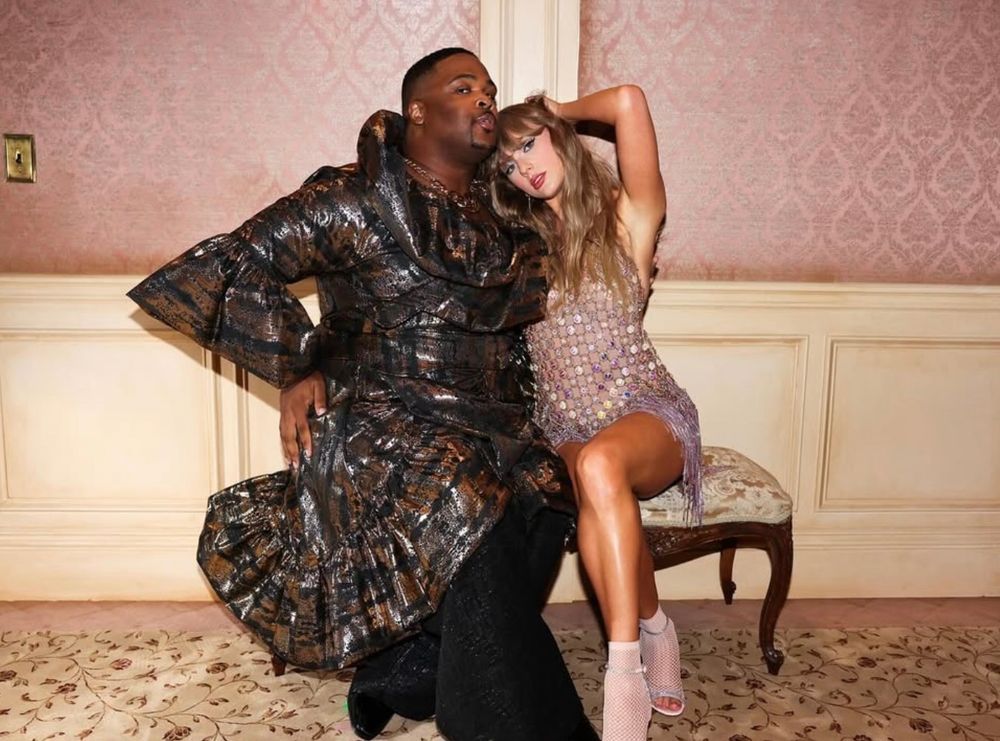 an instagram post by Kam Saunders @kamnsaunders. photo is of cam in a black, bronze and silver metallic jacket and black pants posing on a settee with Taylor Swift who is wearing an iridescent metallic dress, see through metallic peep toe socks and silver sandals. they appear to be in a vintage hotel with creamy printed carpet, cream wainscoting and pink damask wallpaper.