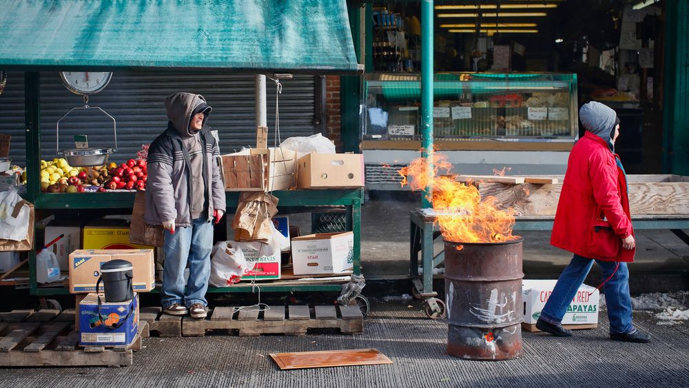 A bustling market scene featuring two persons, one in a grey coat and the other in a red jacket, near a barrel fire, amidst colorful fruit displays.