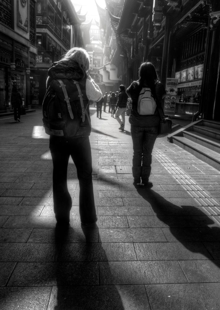 A black and white photograph of a very old looking historic district in Shanghai China. The perspective is from behind two women who are facing away from the viewer, further down the street. Both women are wearing backpacks. The street is paved with stone and the buildings on either side are tall and closely packed together, creating a sense of enclosure. The architecture of the buildings is a traditional Chinese with intricate and overhanging roofs. There are other people visible in the distance, also walking down the street,. The lighting is dramatic, with strong contrasts between light and shadow, which accentuates the textures and shapes of the buildings and the street surface. 