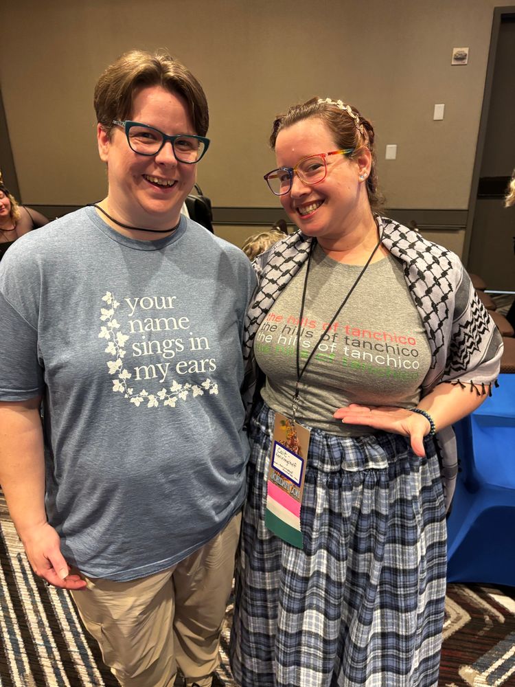 Eilonwy smiling, with glasses, in blue shirt with text “your name sings in my ears”
Cait smiling with glasses in shirt with hills of tanchico text and a keffiyeh
Bigger smiles from both and Cait is emphasizing the hills of Tanchico text with a hand under her chest 