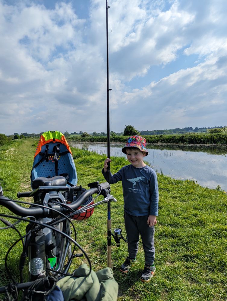 Boy (4) standing on canal bank with a fishing rod, beside longtail e-cargo bike. 