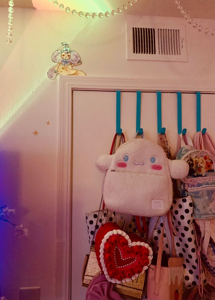 a watercolor paper doll of a purple and yellow plush creature, sitting on top of a door molding. There are cute bags hanging from the door