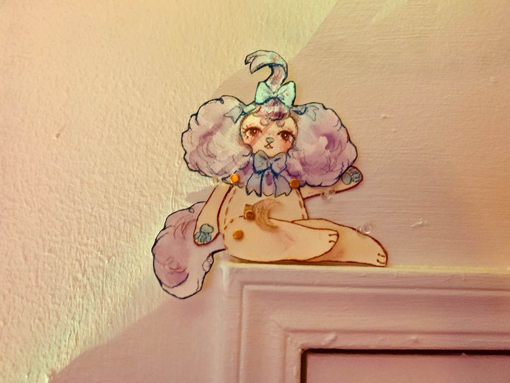 a watercolor paper doll of a purple and yellow plush creature, sitting on top of a door molding.