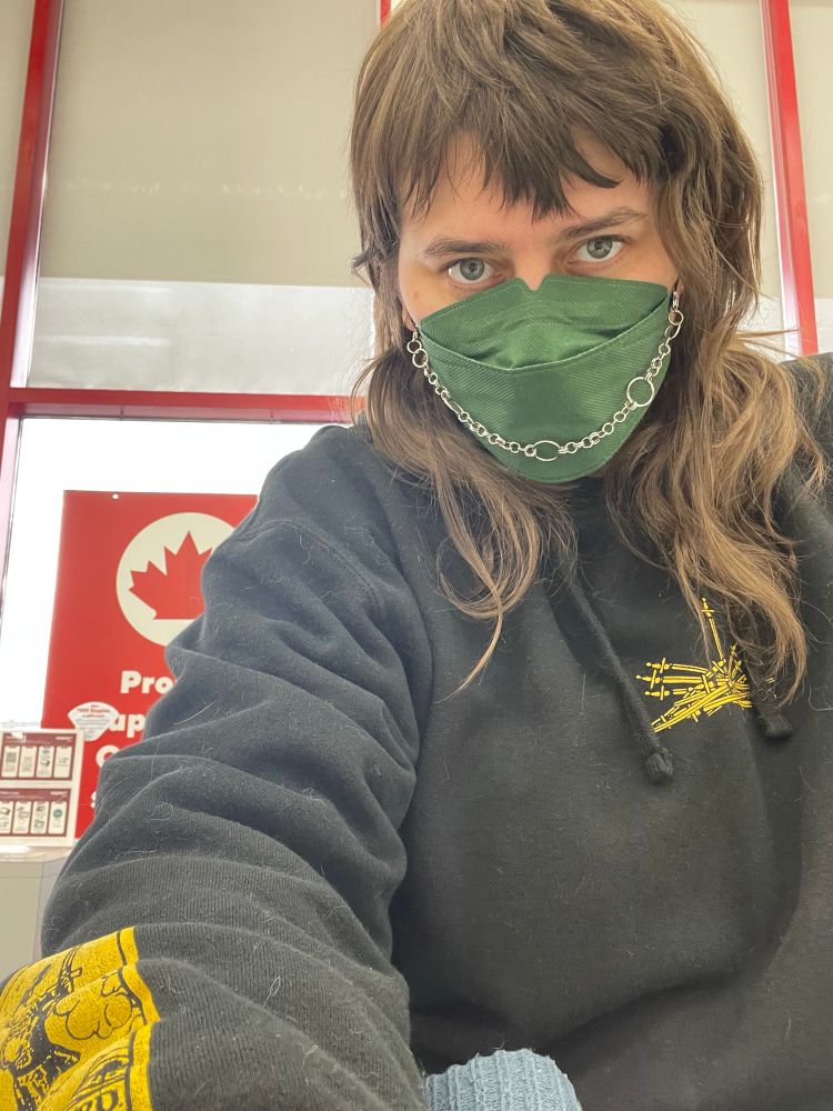 Sid staring down the camera in a green mask with a chain and a hoodie with swords screenprinted on it 