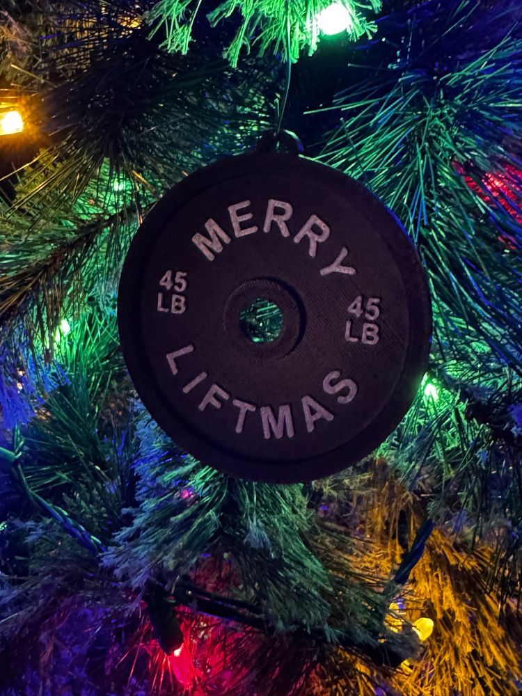 A black weight plate ornament reading "MERRY LIFTMAS" and "45 LB" hanging on a Christmas tree decorated with colorful lights. The tree lights create a festive backdrop of green, blue, red, and yellow illumination around the ornament.​​​​​​​​​​​​​​​​