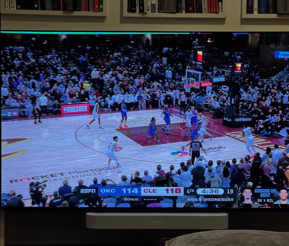 A TV screen showing an NBA basketball game between OKC (Oklahoma City Thunder) and CLE (Cleveland Cavaliers). The score is OKC 114, CLE 118 with 4:36 left in the 4th quarter. The game is being broadcast on ESPN. The TV appears to be mounted below some bookshelves filled with various books, and there's a partial view of what looks like a couch or furniture piece at the bottom of the image.