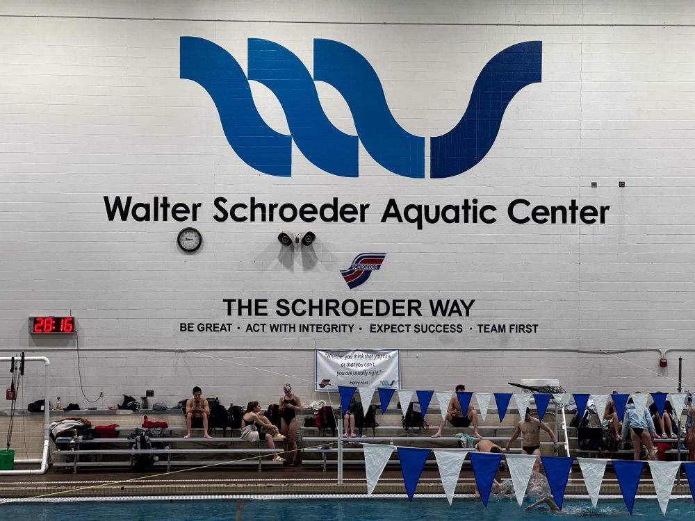 This photo shows the interior of the Walter Schroeder Aquatic Center, featuring its logo on a white wall above the pool. The logo consists of three blue wavelike shapes and the text “Walter Schroeder Aquatic Center.” Below it is the motto, “The Schroeder Way: Be Great • Act With Integrity • Expect Success • Team First.” In the foreground, there are blue-and-white triangular flags hanging above the pool, with swimmers and spectators sitting on benches along the pool’s edge. A digital clock displaying “28:16” is mounted on the wall to the left. The environment suggests an active swimming practice or event.