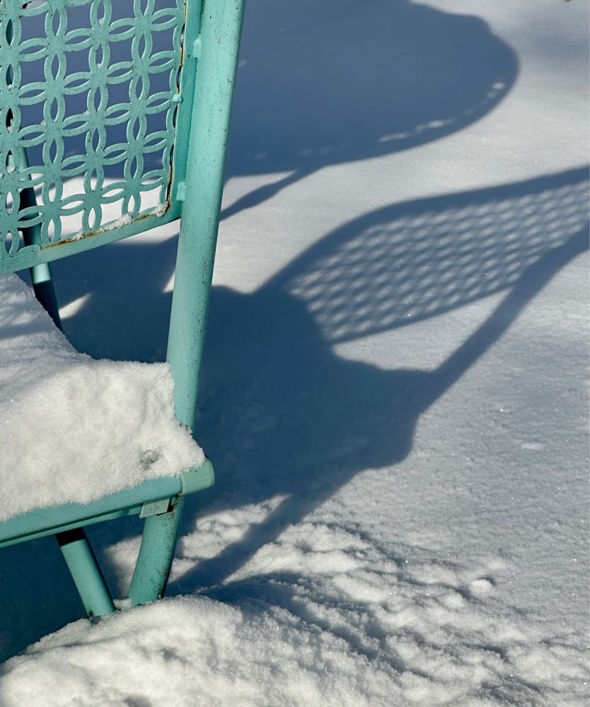 Patterned shadow of a metal chair and round table cast onto fresh white snow in bright sunlight, with a bit of the turquoise chair visible on the left side of the frame. 