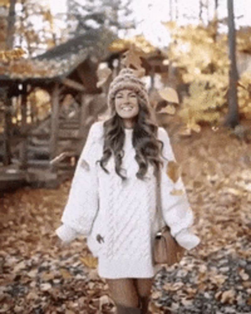 a woman wearing a white sweater dress and a beanie is standing in a pile of leaves .