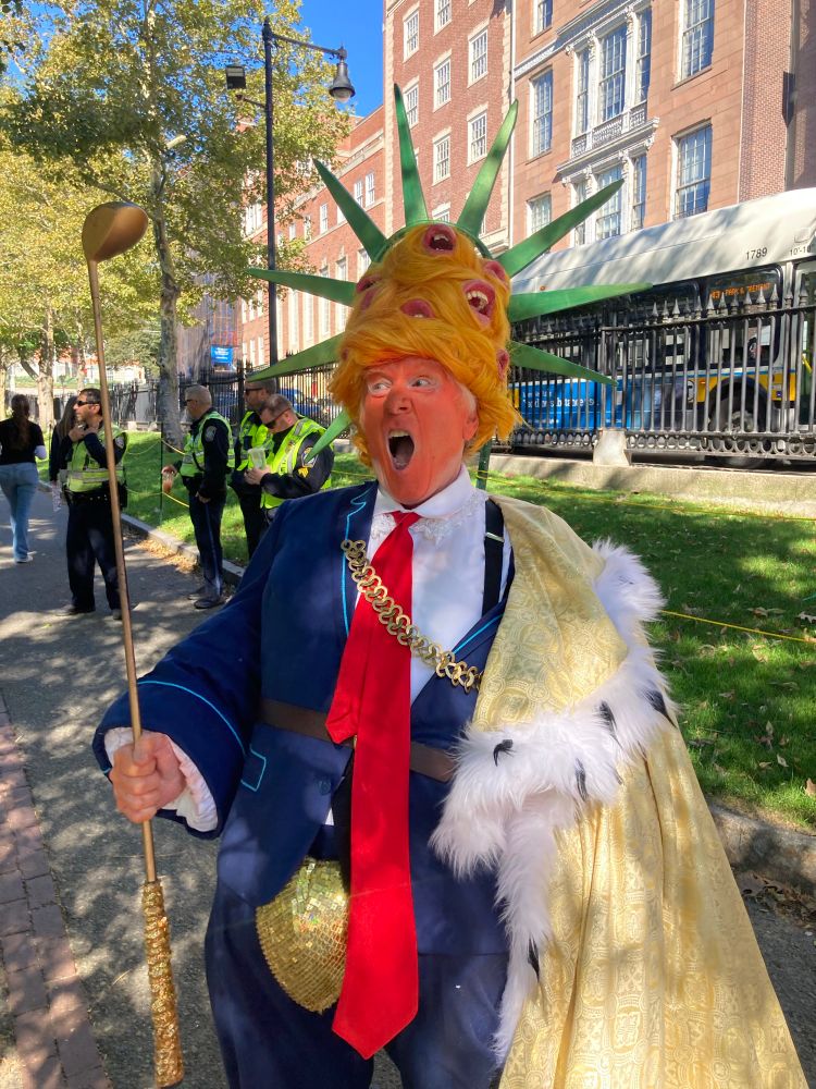 An image of the Mad King Supreme: an orange-faced lunatic in a golden cape, blue suit, red tie, high orange wig filled with horrifying fake human mouths, and a huge sequined codpiece. He holds a golden golf club and his mouth is wide open, as always. 