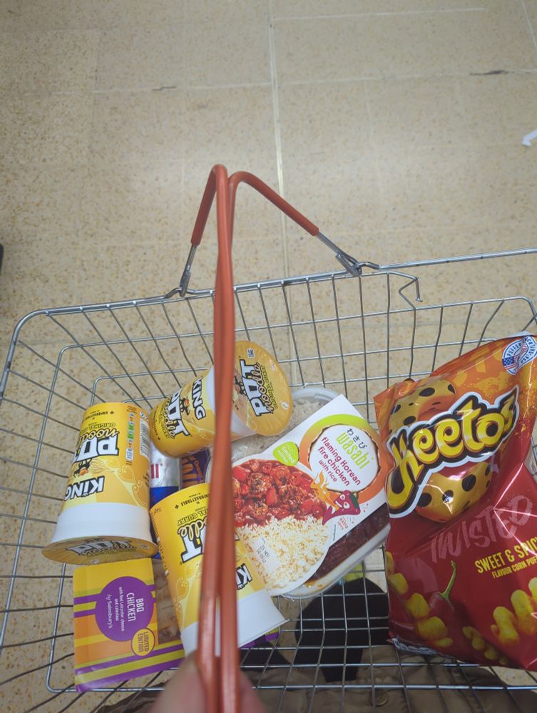A Sainsbury's basket with three king size curry pot noodles, a big pack of Cheetos, a fancy ready meal and a big red bull 