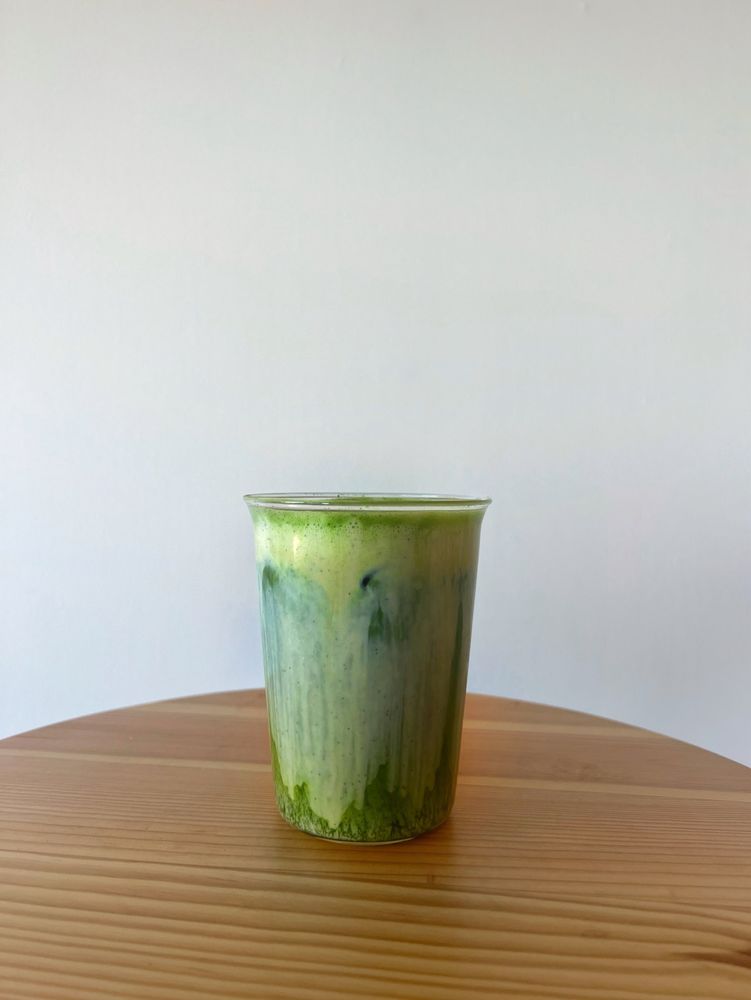 tall glass filled with a gradient of white cream cascading over dark green matcha over ice on top of wood table with white background 