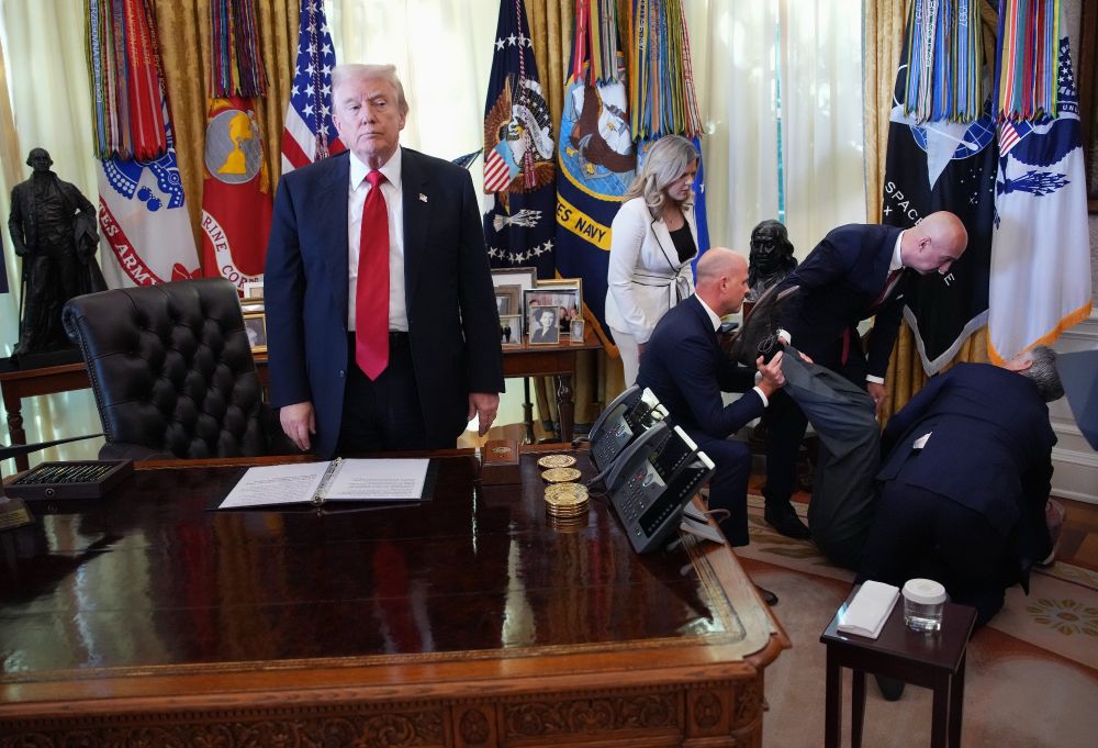 Photo, showing Trump standing alone at the resolute desk and looking forward while several people are several feet to his left, hunched towards the floor, attending to a man who collapsed during a press conference 
