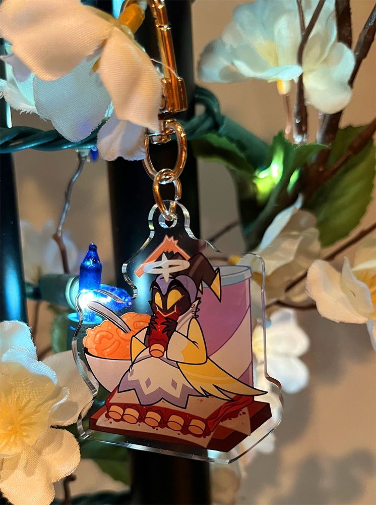 Keychain  Adam from Hazbin Hotel. He's sitting and eating a giant rack of ribs with a drink in a cup and a bowl of mac n cheese behind him.