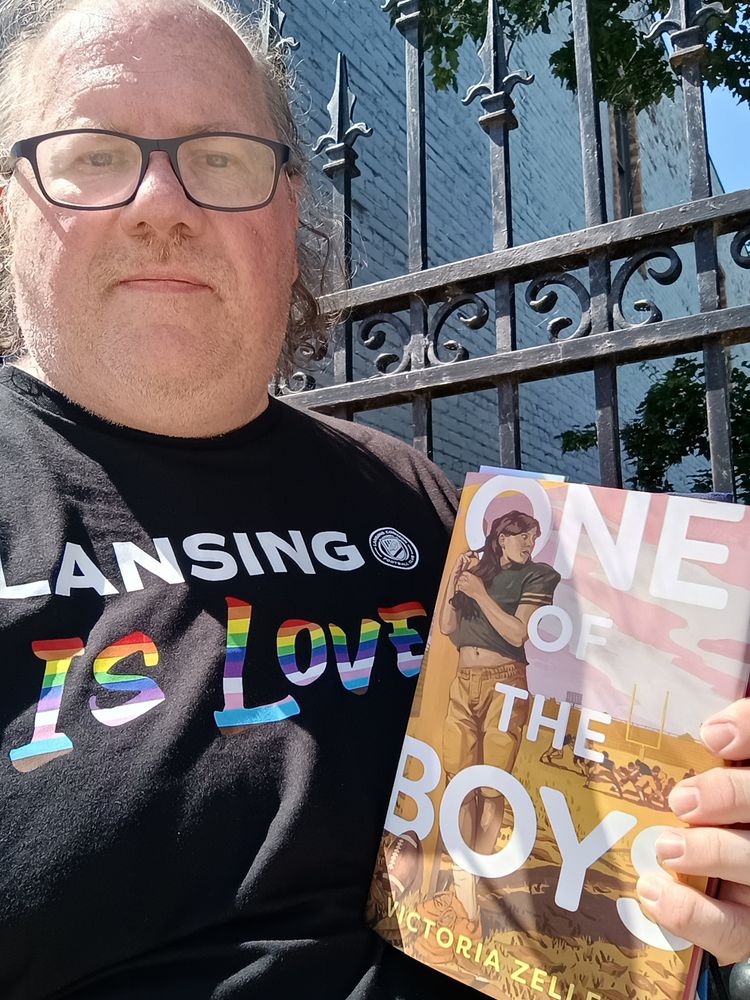 "One of the Boys" by Victoria Zeller.  40-something guy in a "Lansing is Love" tshirt holding the book pleased with the purchase even if I took this too quickly to get a big smile in
