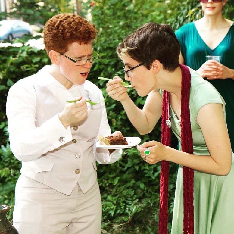 Two people eating pie on their wedding day. The person on the right just took a bite of pie off her own fork, the person on the left is aghast