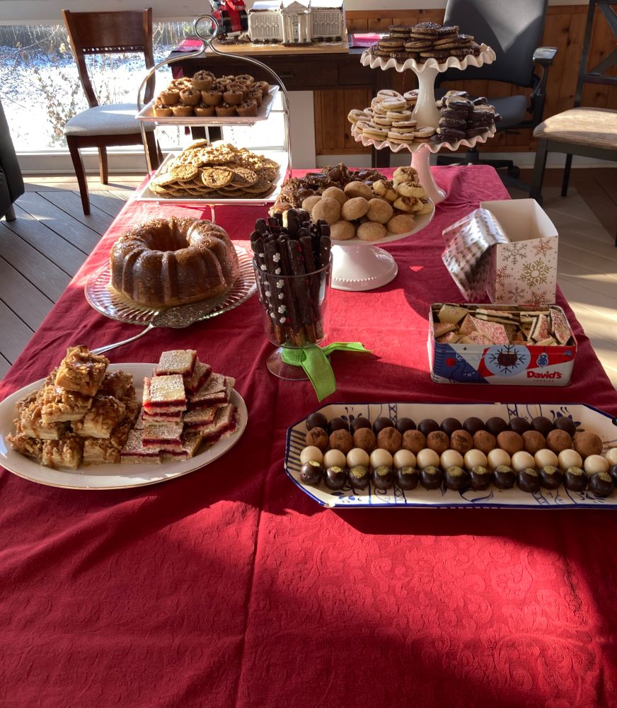 Table filled with homemade treats. Clockwise from lower left: salted caramel apple pie bars, lemon cranberry bars, Irish crème Bundt cake, molasses spice, white chocolate macadamia oatmeal, maple pecan, peanut butter cups with mascarpone chocolate ganache, hazelnut espresso sandwiches, Linzer with raspberry and pistachio, orange shortbread, turtle thumbprints, baklava thumbprints, snickerdoodles, chocolate dipped pretzel rods, peppermint bark, four kinds of truffles (pistachio, lemon, tiramisu, bourbon). Plus gingerbread White House in background