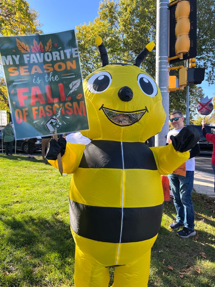 Protester in a bee costume with sign “my favorite season is the fall of fascism”