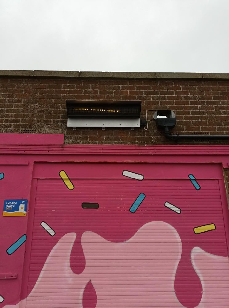 "A pink roller shutter door decorated with colourful sprinkles and dripping patterns, set in a brick building. Above the door are a light fixture and a security camera. A small blue sign to the left reads 'Seaside Award Hours'. The sky is overcast."