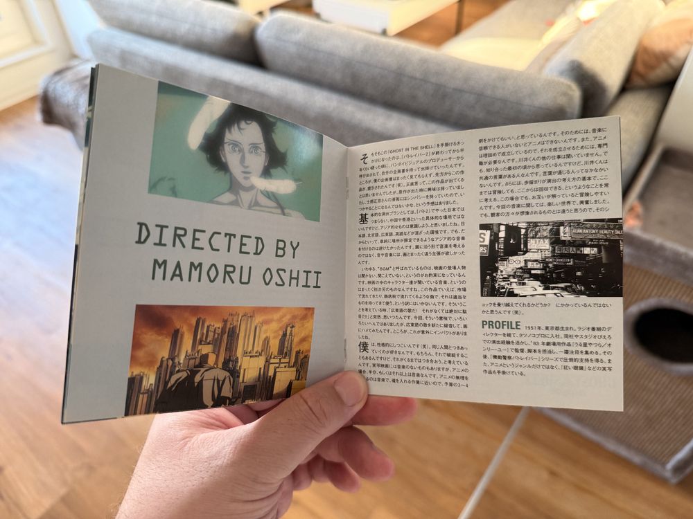 Ein geöffnetes CD-Booklet zum Anime-Film „Ghost in the Shell“. Links steht groß „Directed by Mamoru Oshii“ unter einem Porträt der Hauptfigur Motoko Kusanagi und einem Bild einer futuristischen Stadt. Die rechte Seite zeigt einen langen Textblock auf Japanisch mit dem Abschnitt „PROFILE“, der Informationen zu Mamoru Oshii enthält. 
