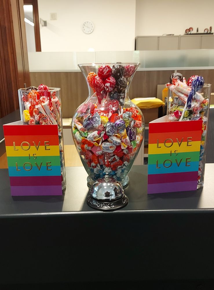 Three vases full of individually wrapped candies of many colours are flanked by two rainbow cards reading "Love is Love." 