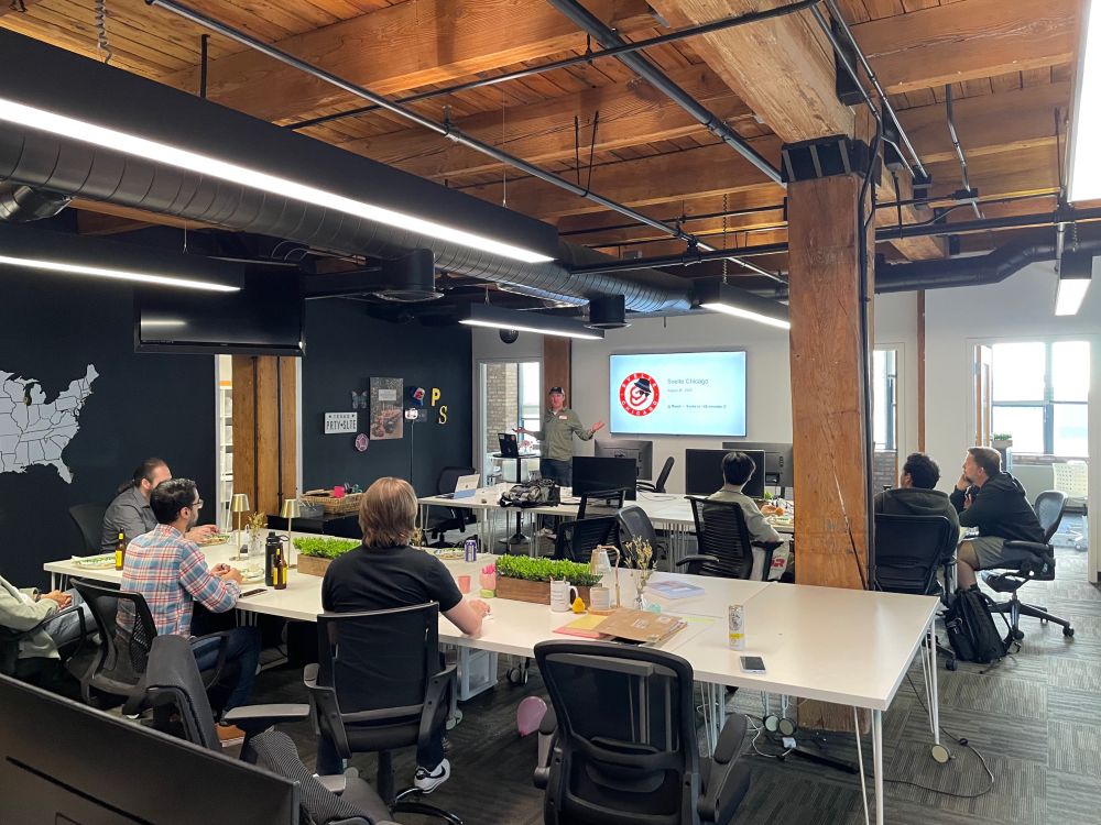 Speaker presenting at Svelte Chicago meetup in modern office space. Attendees at conference tables with pizza, watching presentation on screen.