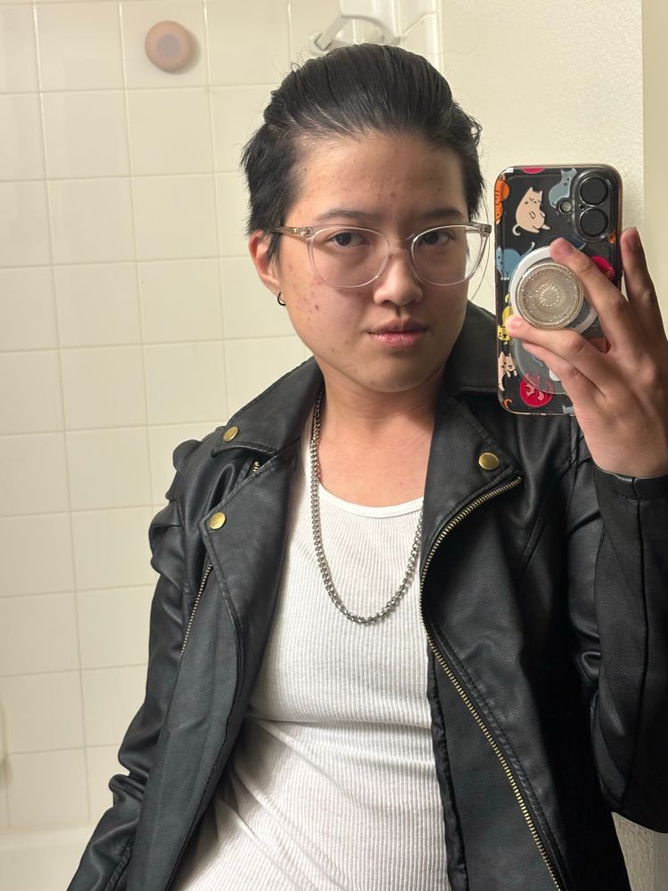 Noah with hair pulled back— dressed in a white tank top, leather jacket, and silver chain necklace 