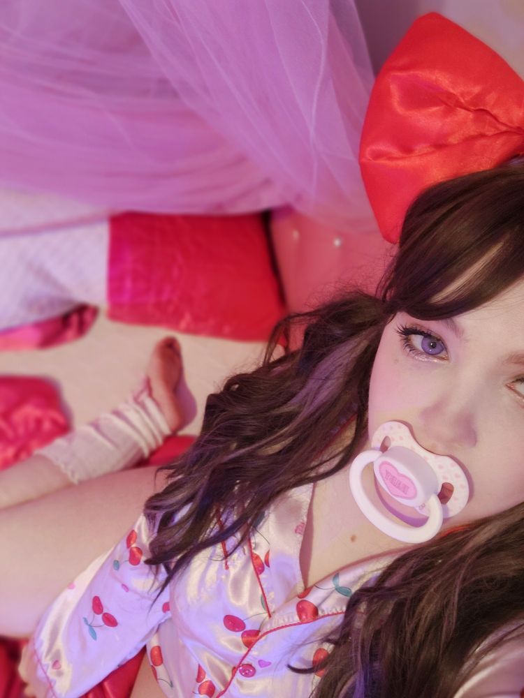 Aeris is wearing a large red bow on her head. Her thigh and calf with bow leg warmers are visible in the distant background. She's wearing red and pink cherry print pajamas with brunette pigtails. Her eyes are purple colored. Her aegyo sal are highlighted with glitter.