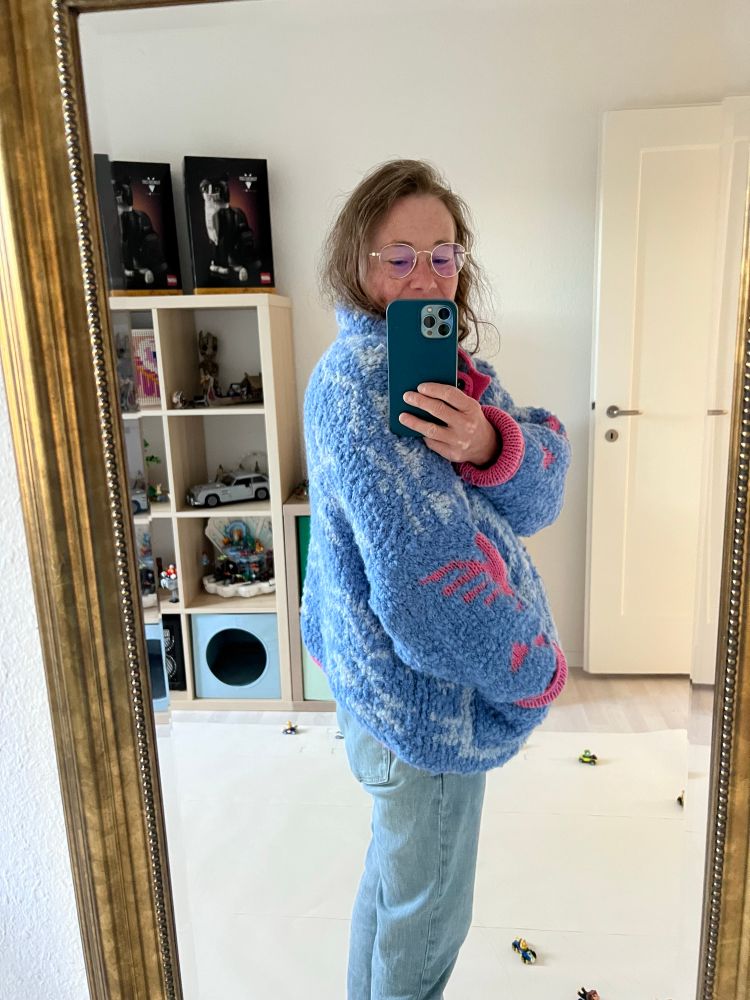 Eine Frau trägt eine selbstgestrickte Jacke und steht seitlich vor einem Spiegel. Die Grundfarbe der Jacke ist blau, das Muster hellblau und rosa. Im Hintergrund sieht man verschiedene Lego Sets. 
