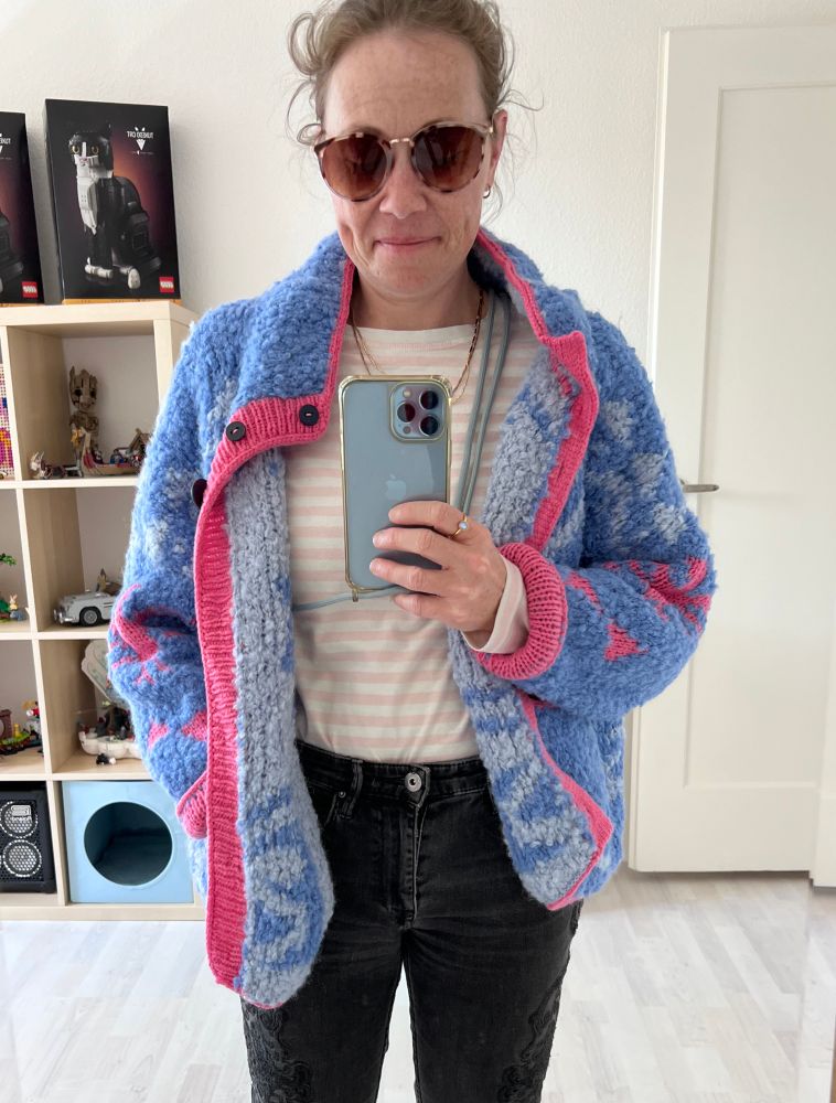 Ich trage eine Strickjacke in hellblau, dunkelblau und pink. Sie hat verschiedene Muster und ist aus flauschiger Wolle gestrickt