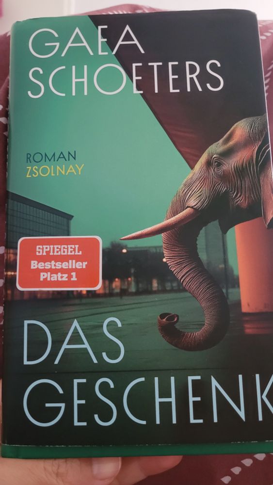 Foto eines Buchcovers ' Das Geschenk' von Gaea Schoeters. Aufkleber: Piegel Bestseller Platz 1. Man sieht einen grossen Elefantenkopf auf einen grossen leeren Stadtplatz blicken 