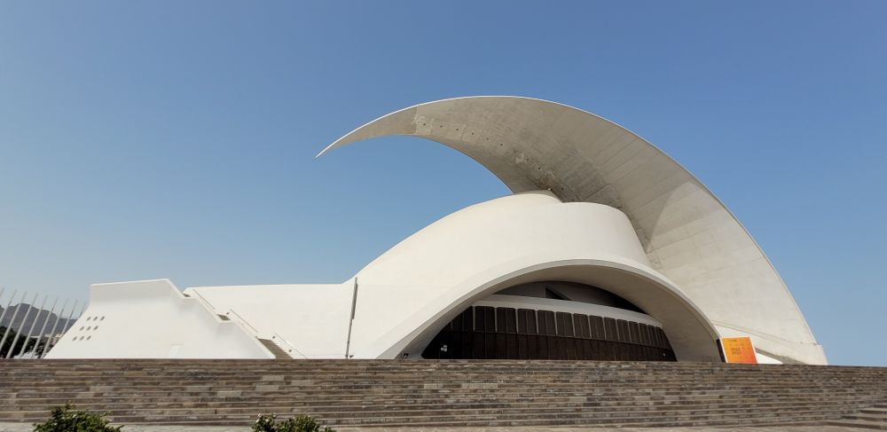 Viele Treppen führen hoch zu einem länglichen Gebäude. Das Gebäude ist mit weissen Fliesen überzogen. Von rechts nach links sieht man eine geschwungene Betonplatte hoch über das Dach ragen, sie sieht aus wie eine Schwanzflosse.