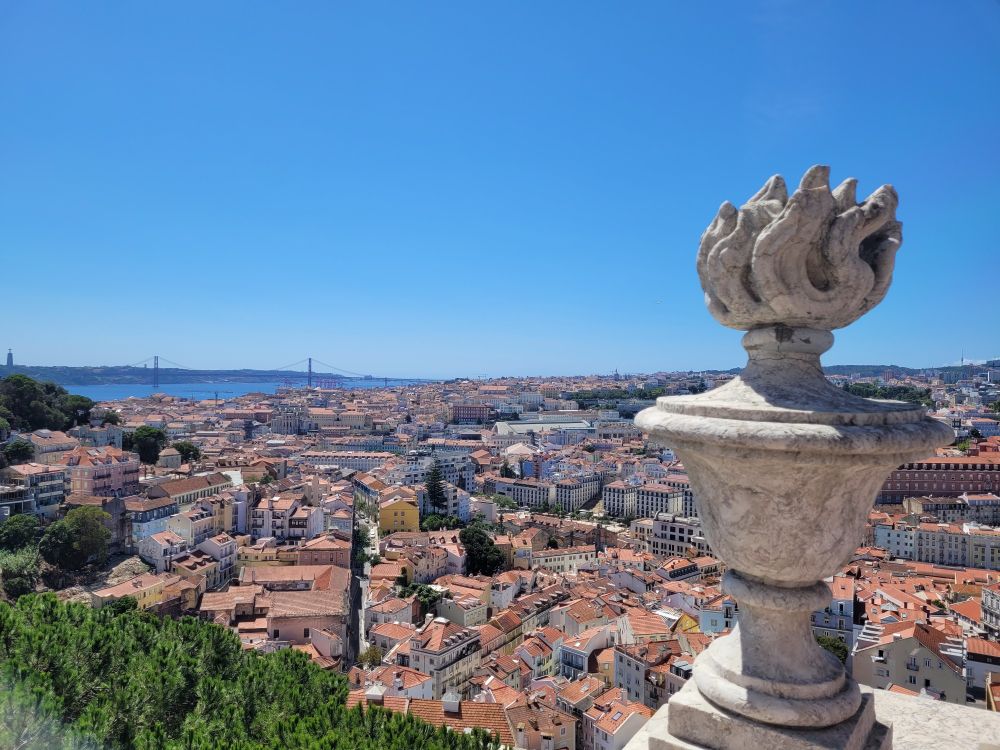 Blick von einem Turm auf Lissabon. Man sieht viele Häuser mit roten Dächern und enge Gassen. Im Vordergrund ein Stück Mauer, darauf an der Ecke eine Skulptur aus Stein, eine Fackel. Im Hintergrund der Fluss Tejo und die Brücke des 25. Aprils.