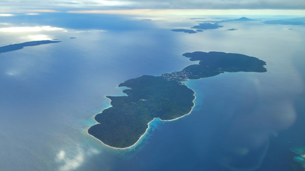 Eine kleine längliche Insel mit vielen Buchten von einem Flugzeig aus fotografiert. Die Buchten schimmern türkis im Hintergrund weitere Inseln, die aus dem Wasser ragen.